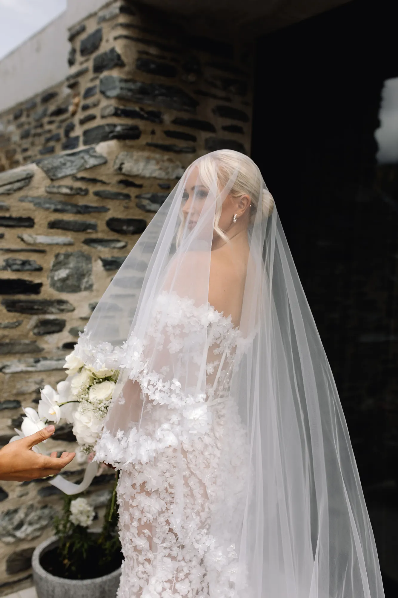 Platinum low bun under a long cathedral veil, against a schist stone wall